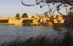 an old village sits on the edge of a body of water with tall grass in front of it