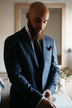 a bald man in a suit and tie is looking down at his watch while standing next to a bed