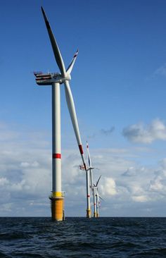 several wind turbines in the ocean with blue sky and clouds behind them, all connected to one another