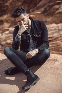 a man sitting on top of a rock with his hand to his face while wearing black boots