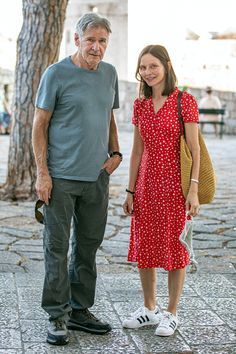 an older man and woman standing next to each other in front of a tree on the sidewalk