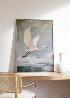 a large white bird flying over a wooden table next to a chair and painting on the wall