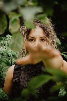 a woman with curly hair is looking at the camera through some trees and leaves while holding her hand up in front of her face