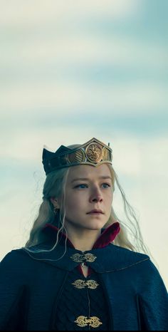 a woman wearing a crown and cape with long blonde hair standing in front of a cloudy sky