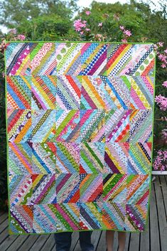 a woman holding up a colorful quilt on top of a wooden deck with flowers in the background