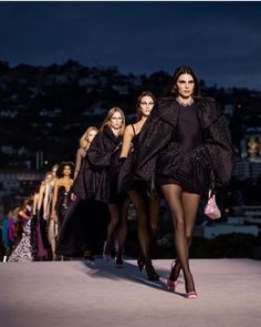 models walk down the runway in black dresses and high heeled shoes, with city lights behind them