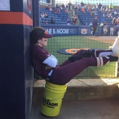 a woman sitting on top of a bucket in front of a baseball field