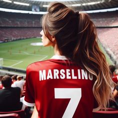 a woman in a red soccer jersey looks out at the field from behind her is a stadium full of people