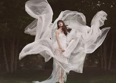 a woman is standing in the grass with her dress flowing around her and there are trees behind her