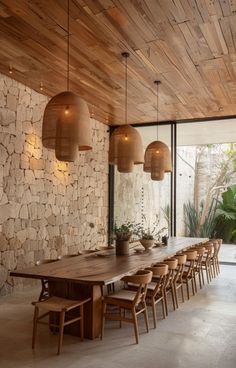 a wooden table surrounded by chairs and lights in front of a stone wall with large windows