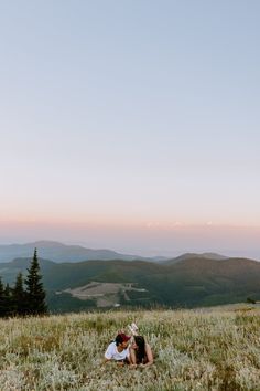 two people laying in the grass on top of a hill with mountains in the background