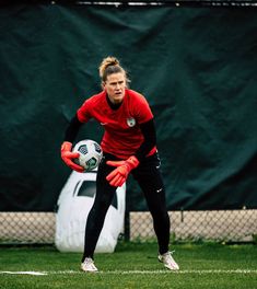 a woman in red shirt and black pants holding a soccer ball on grass with fence behind her