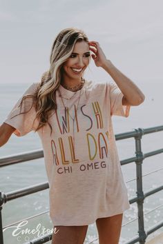 a woman standing on a pier next to the ocean wearing a pink wish all day t - shirt