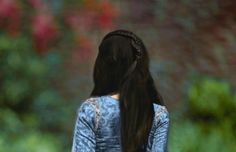 a woman with long black hair standing in front of some trees and bushes looking at something