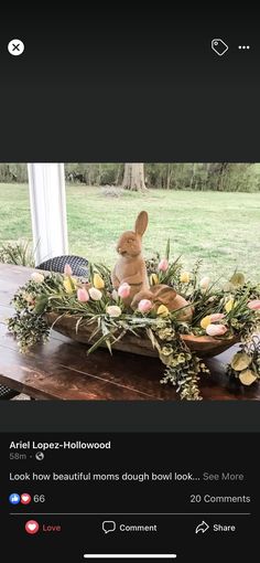 a rabbit sitting on top of a wooden table next to flowers and greenery in front of a window