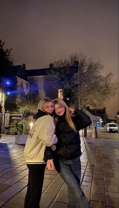 two women standing next to each other in front of a building at night with their arms around each other