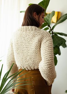 a woman standing in front of a potted plant wearing a white sweater and brown pants