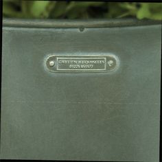 a close up of a metal planter with plants in the background and a name plate on it