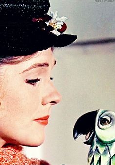 a woman wearing a black hat and holding a green parrot on her shoulder, with another bird in the background