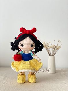 a crocheted doll with a red hat and yellow dress is standing next to a white vase