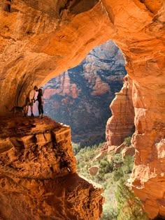 two people standing on the edge of a cliff in front of a canyon with mountains and trees