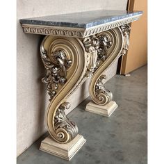 an ornate console table with marble top and gold leaf design on the base, in front of a beige stucco wall