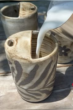 a white pitcher pouring milk into two ceramic vases on a wooden table next to water