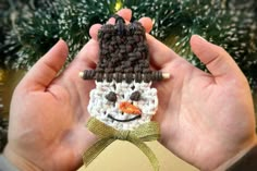 a hand holding a crocheted snowman ornament in front of a christmas tree