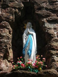 a statue of the virgin mary in front of a rock wall with flowers growing out of it