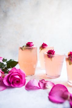 three glasses filled with liquid and flowers on a white tablecloth next to pink roses