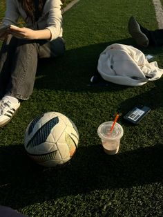 a woman sitting on the grass next to a soccer ball and a cup with a straw in it