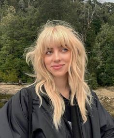 a woman with long blonde hair standing in front of some trees and water wearing a black jacket