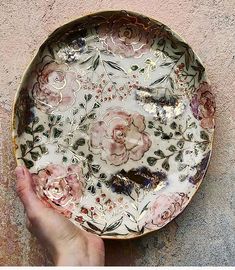 a hand holding a decorative plate with flowers on it and gold trimmings around the edge