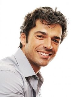 a man with curly hair smiling at the camera and wearing a gray shirt, white collared shirt and black pants