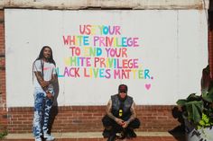 two young men sitting on the ground in front of a wall with words painted on it