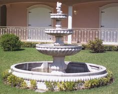 a fountain in the middle of a lawn with bushes around it and a building behind it