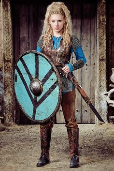 a woman dressed in medieval clothing holding a shield and two swords, standing next to a wooden door