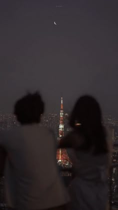 two people are looking at the moon in the night sky over a cityscape