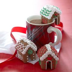 three gingerbread houses sitting next to a cup of hot chocolate on a red table