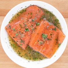 two pieces of cooked salmon are in a white bowl on a wooden table with herbs and seasoning