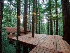 a wooden deck in the middle of a forest with lights strung from it's sides