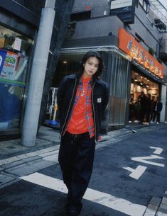 a young man walking down the street in front of a store with an orange shirt and black pants