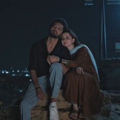 a man and woman sitting on top of a rock in front of a city at night