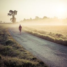 a man walking down a road in the middle of a foggy field at sunset