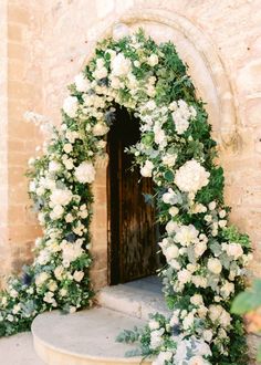 an arch covered in white flowers and greenery next to a doorway with a wooden door