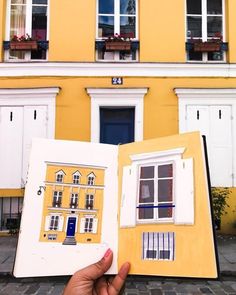 a hand holding an open book in front of a building with windows and shutters