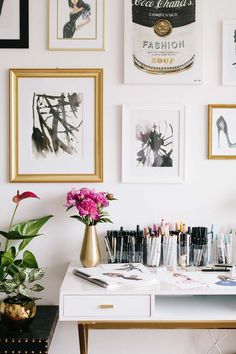 a white desk topped with lots of art next to a wall filled with pictures and flowers