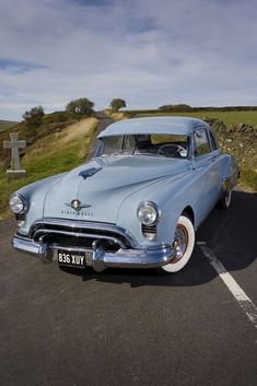 an old blue car is parked in a parking lot next to a stone wall and grass