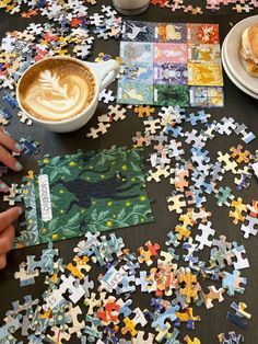 a table topped with lots of puzzle pieces next to a cup of cappuccino