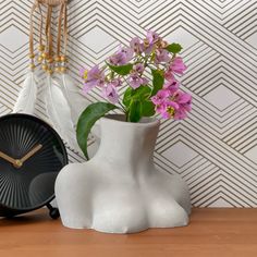 a white vase with purple flowers in it next to a clock on a wooden table